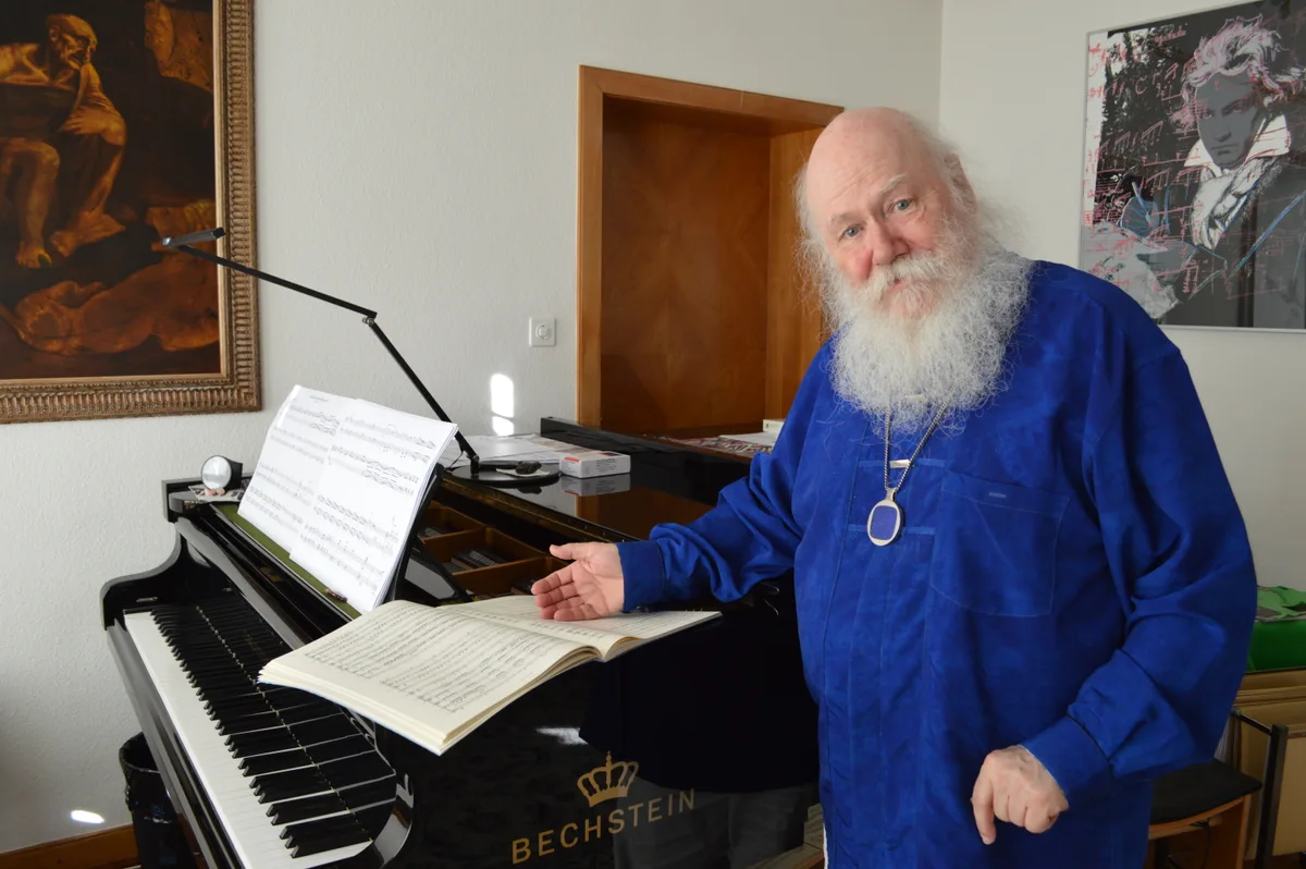 Ein Mann in Blau steht vor dem Klavier und zeigt auf eine Partitur.